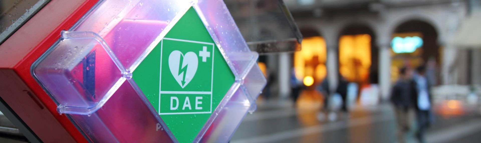 An automated external defibrillator placed outdoors in a transparent case with green signage, located in an urban square with historic buildings in the background and people walking. An automated external defibrillator placed outdoors in a transparent case with green signage, located in an urban square with historic buildings in the background and people walking.