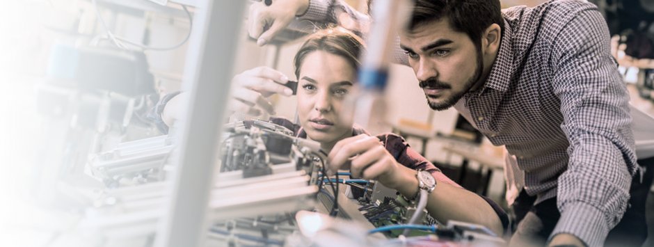 Due persone, una donna e un uomo, lavorano insieme su un macchinario tecnico in laboratorio, concentrati su componenti elettronici e meccanici.