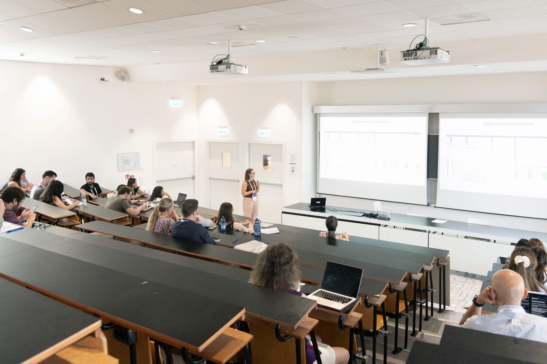 Un'aula con persone sedute ai banchi mentre un uomo presenta di fronte a loro.