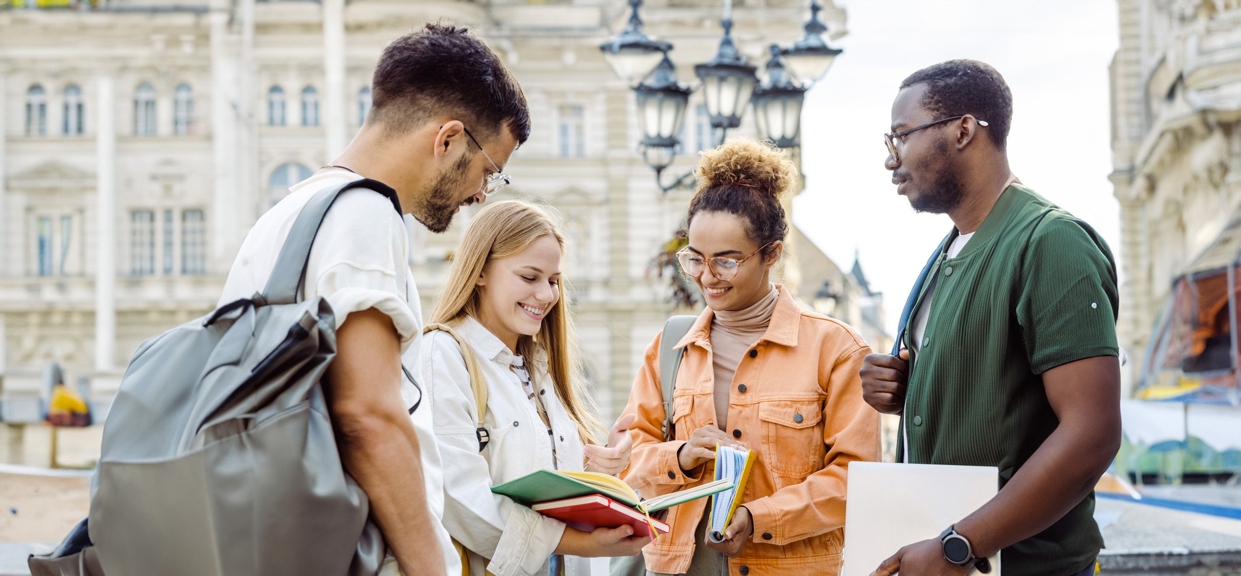 Group of four international students talking and smiling outdoors in a European square, with historic buildings in the background.