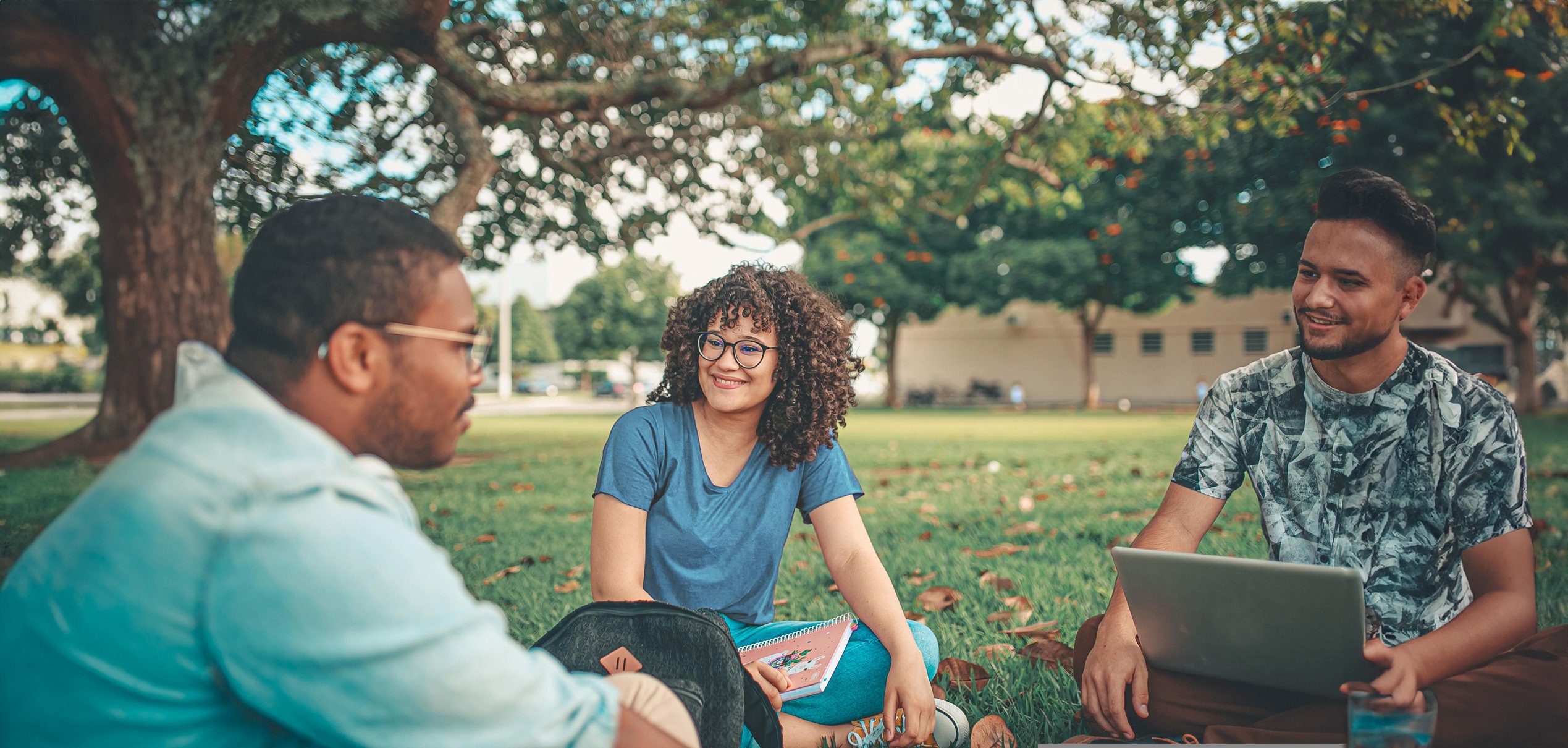 Tre studenti seduti sull'erba con laptop, impegnati in attività di studio e collaborazione.