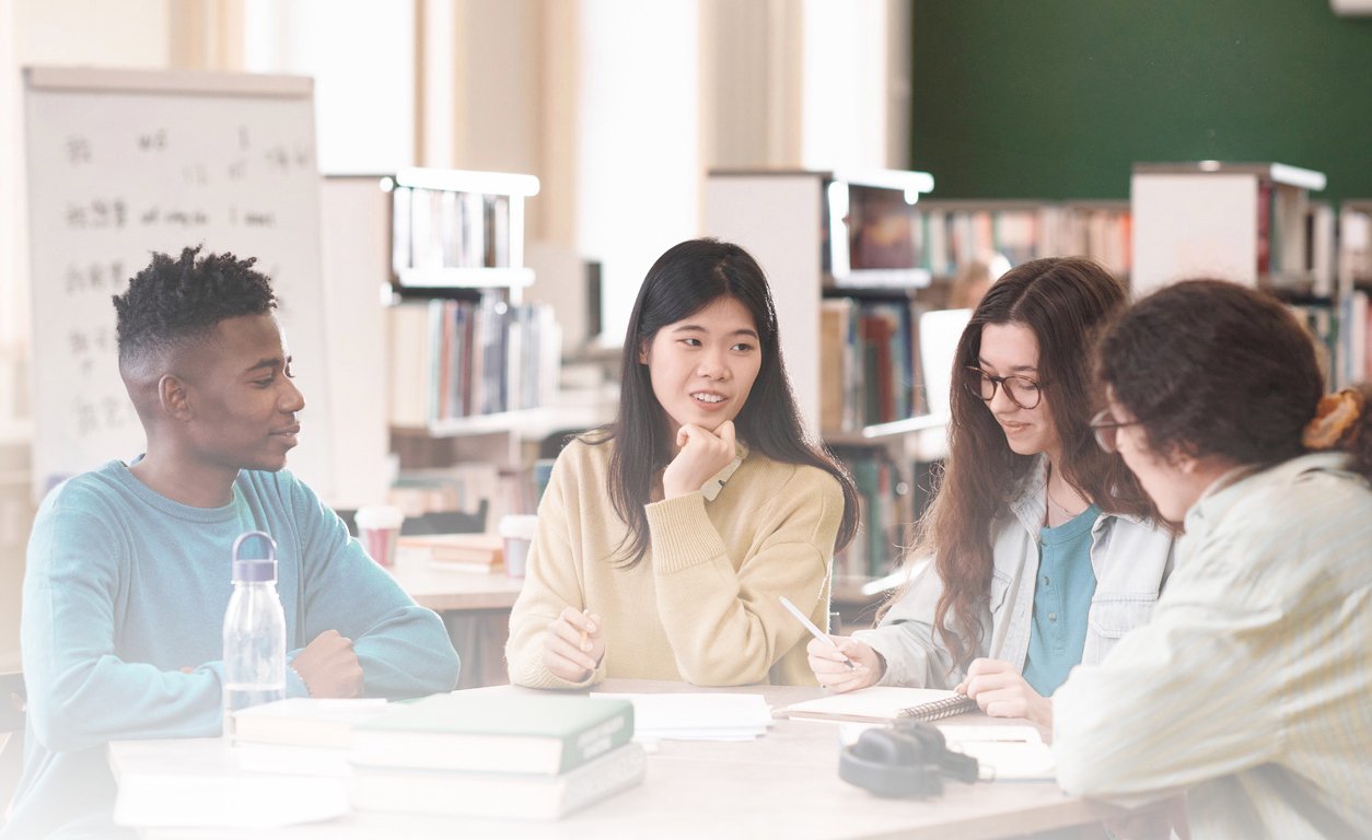 Gruppo di giovani studenti di diverse etnie che discutono e scambiano idee in una biblioteca o ambiente di studio.